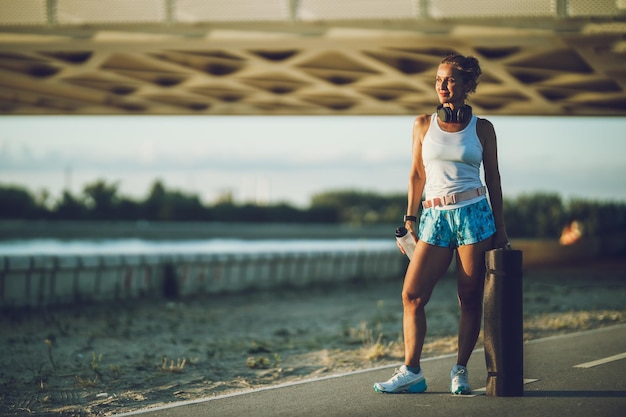 Mujer en forma de mediana edad con colchoneta de yoga y botella de agua está descansando cerca del río en la ciudad antes de un entrenamiento duro.