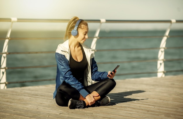 Mujer en forma relajada en ropa deportiva con auriculares elige música de un teléfono inteligente para entrenar en la playa en un día soleado