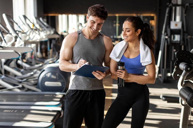 Una mujer en forma con una toalla en los hombros hablando con su entrenador masculino durante el entrenamiento en el gimnasio discutiendo el plan