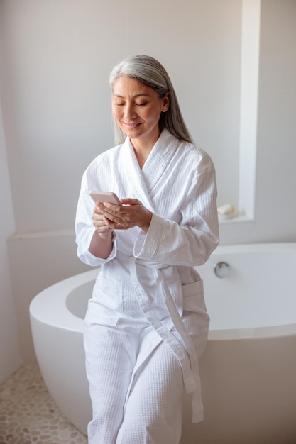 Mujer de foto vertical sentada en el borde de la bañera usando un teléfono inteligente