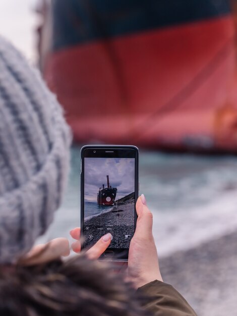 Una mujer fotografía un gran barco amarrado a la orilla en su teléfono celular mientras camina por la playa