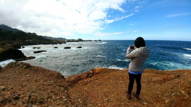 Foto mujer fotografiando el mar mientras está de pie en una formación rocosa contra el cielo