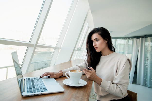 Mujer freelance trabajando en una computadora portátil y escribiendo en el teléfono inteligente al cliente