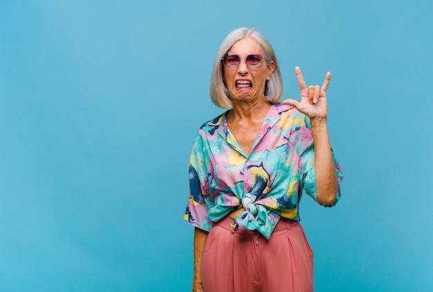 Mujer fresca de mediana edad que se siente feliz, divertida, segura, positiva y rebelde, haciendo un letrero de rock o heavy metal con la mano