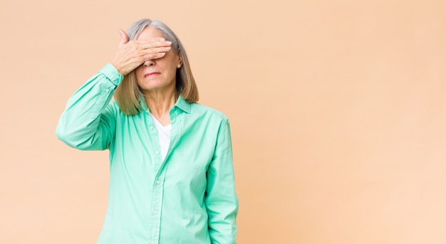 Mujer fría de mediana edad que cubre los ojos con una mano sintiéndose asustada o ansiosa, preguntándose o esperando ciegamente una sorpresa