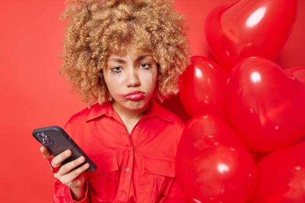 Una mujer frustrada e infeliz con el pelo rizado y tupido ha estropeado el maquillaje sostiene el teléfono móvil y un montón de globos de helio del corazón usa una camisa aislada sobre fondo rojo espera a su novio el día de San Valentín