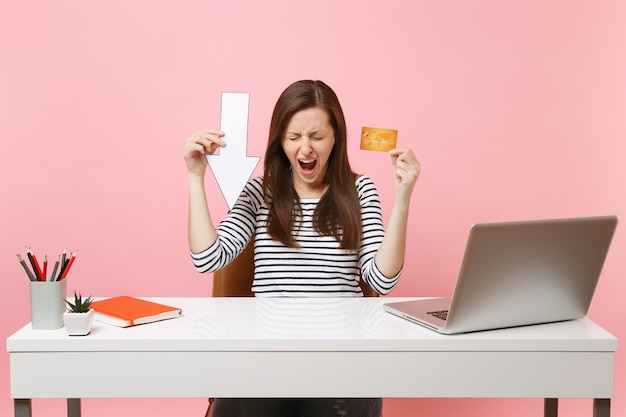 Mujer frustrada gritando sosteniendo el valor caída flecha tarjeta de crédito sentarse y trabajar en el escritorio blanco con computadora portátil contemporánea