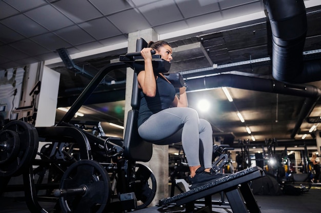 Una mujer fuerte está haciendo ejercicios para piernas y hombros en una máquina de ejercicios en un gimnasio