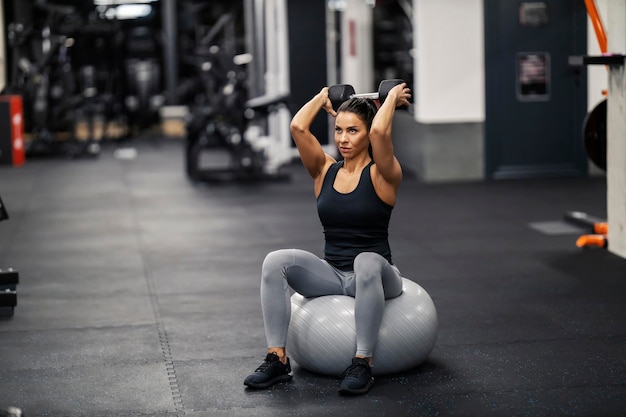 Una mujer fuerte está sentada en una pelota de pilates en un gimnasio y haciendo ejercicios de fuerza para bíceps