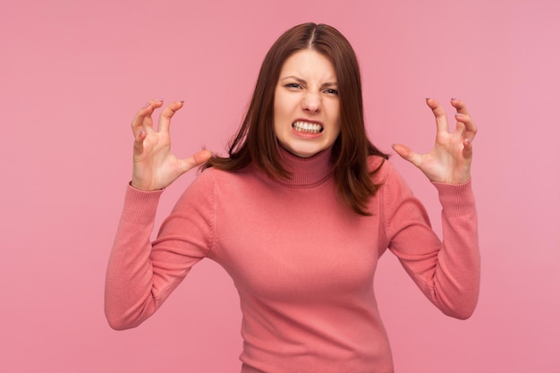 Mujer furiosa extremadamente enojada con el pelo castaño haciendo muecas y apretando los dientes mirando a la cámara con los brazos levantados, depresión. Disparo de estudio interior aislado sobre fondo rosa