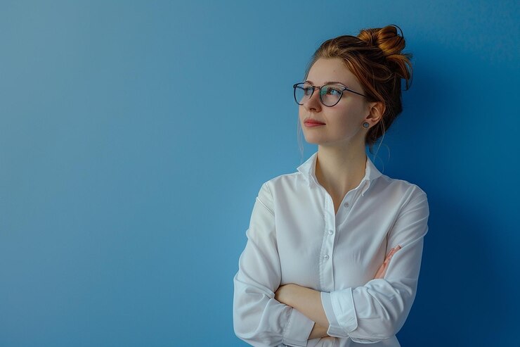 una mujer con gafas y una camisa blanca de pie contra una pared azul