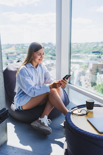 Mujer con gafas clásicas escuchando música con auriculares desde el teléfono y sonriendo mientras se sienta en un moderno espacio de coworking mujer milenaria despreocupada con gafas disfrutando del tiempo libre para comunicarse