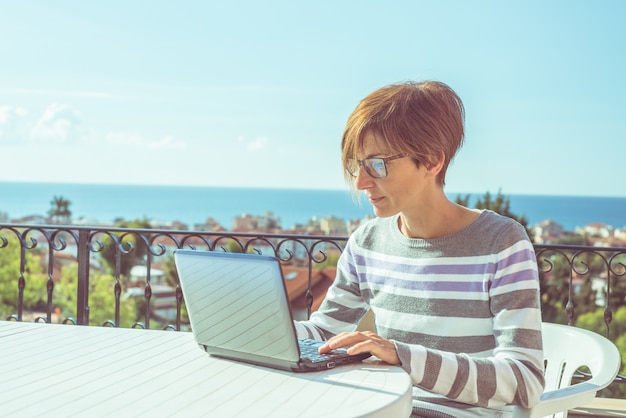 Mujer con gafas y ropa casual trabajando en la computadora portátil al aire libre en la terraza