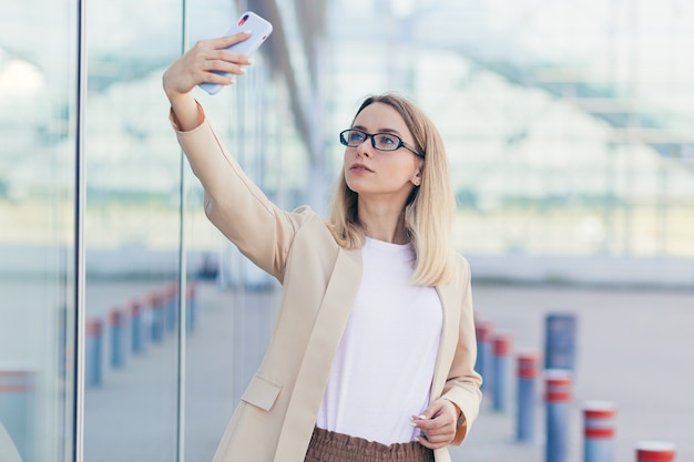 Mujer con gafas rubia sosteniendo un teléfono móvil toma una foto selfie