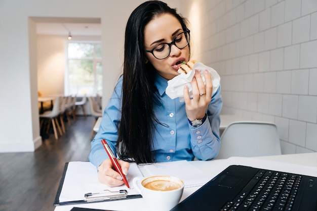 Una mujer con gafas se sienta en una mesa en la oficina y come un sándwich