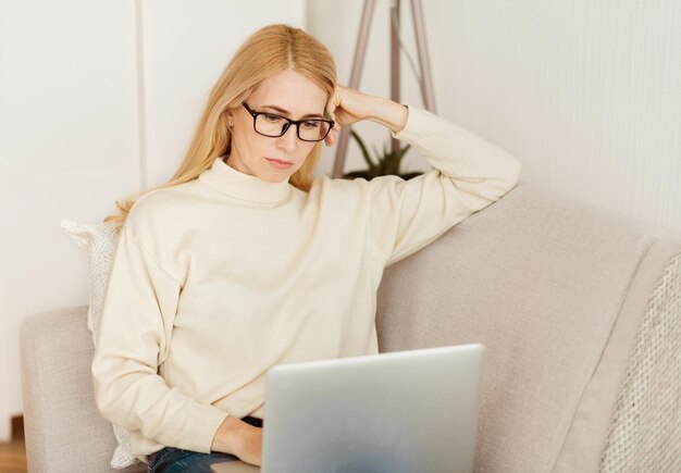 Una mujer con gafas se sienta en un sofá trabajando en una computadora portátil está vestida con un suéter blanco y