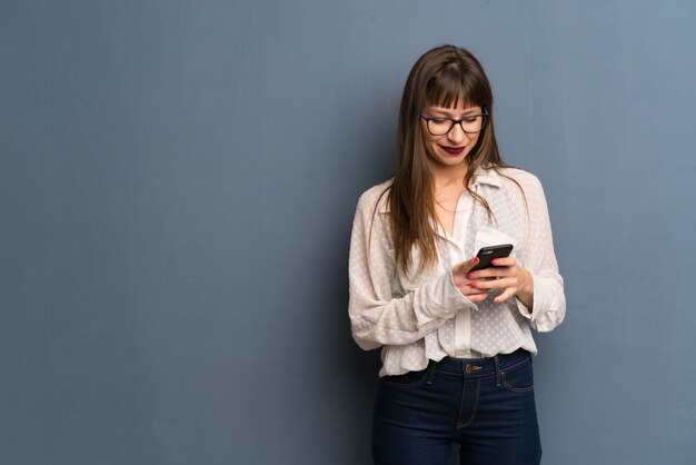 Foto mujer con gafas sobre muro azul enviando un mensaje con el móvil.