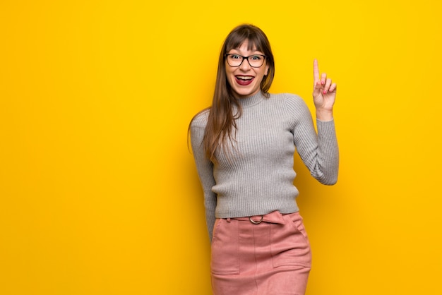 Mujer con gafas sobre una pared amarilla con la intención de realizar la solución mientras levanta un dedo.