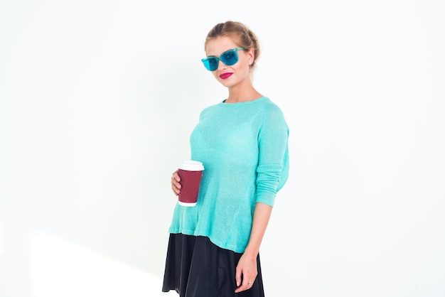 Mujer con gafas de sol azul menta y top con peinado desordenado sonriendo Retrato de modelo femenino glamoroso rubio en forma posando sosteniendo una taza de café roja sobre fondo blanco no aislado