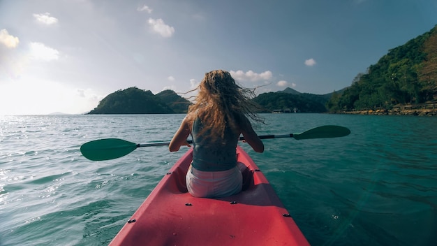 Mujer con gafas de sol filas canoa de plástico rosa a lo largo de agua de mar contra verdes colinas y cielo azul