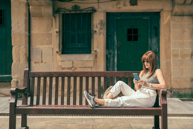 Mujer con gafas de sol sentada en un banco de madera usando un teléfono celular en un antiguo pueblo