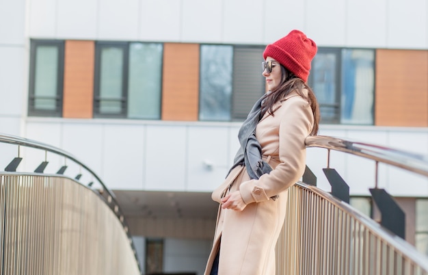 Mujer con gafas de sol, sombrero rojo y abrigo de pie sobre el puente en la ciudad de otoño.