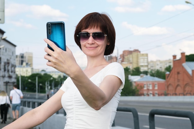 Mujer con gafas de sol sonríe a la pantalla de su teléfono inteligente en su mano extendida para tomarse una foto en el teléfono