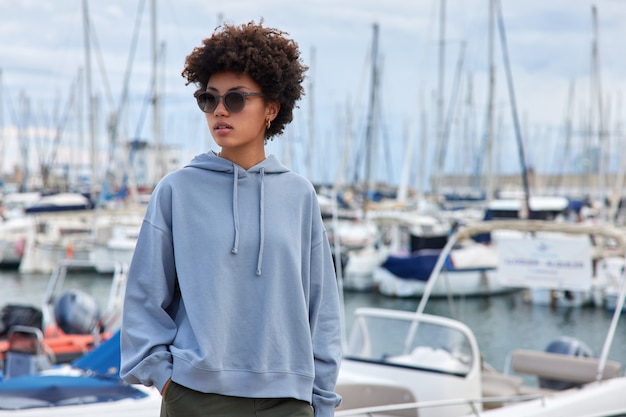 Foto mujer con gafas de sol y sudadera con capucha se encuentra en el puerto durante el día soleado disfruta de hermosos paisajes paseos al aire libre para ir a hacer una excursión por el mar en yate