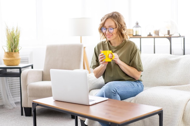 Mujer con gafas usa el teclado de mecanografía portátil sentado sofá gran ventana fondo interior de la casa Mujer independiente trabajando desde casa Estudiante de aprendizaje a distancia relajante ver lecciones video conferencia