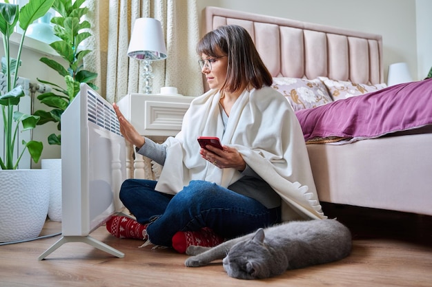 Foto mujer con gato de mascota en casa calentándose cerca de un calentador eléctrico mujer de mediana edad descansando en el suelo usando teléfono inteligente dormitorio de calefacción con radiador temporada fría otoño invierno comodidad crisis energética