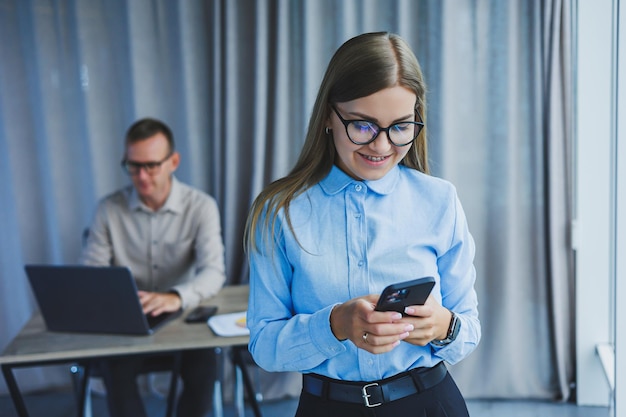 Mujer gerente con anteojos clásicos sonriendo durante las horas de trabajo en la oficina hablando por teléfono con colegas portátiles en el fondo Un colega está en el enfoque selectivo de fondo