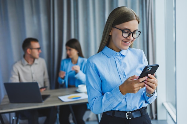 Mujer gerente con anteojos clásicos sonriendo durante las horas de trabajo en la oficina hablando por teléfono con colegas portátiles en el fondo Un colega está en el enfoque selectivo de fondo