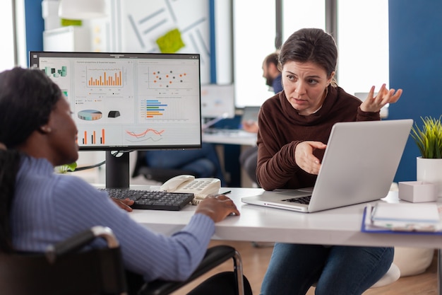 mujer gerente con máscara trabajando con documentos financieros comprobando gráficos, compañero de trabajo apuntando a la computadora portátil, leyendo informes