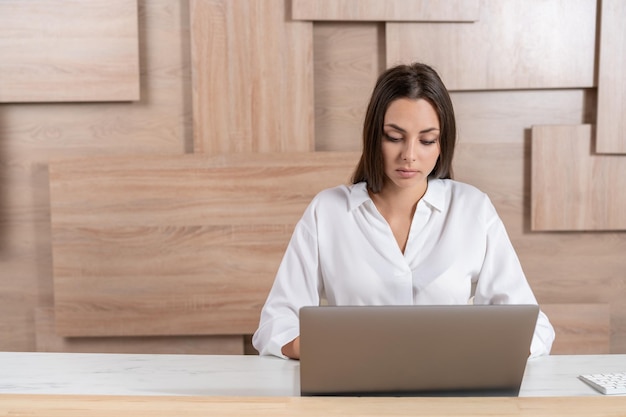 Mujer gerente de oficina con camisa blanca sentada a la mesa con una computadora portátil en el fondo de la pared de la oficina de madera Trabajador de oficina concentrado mirando la pantalla Concepto de trabajo
