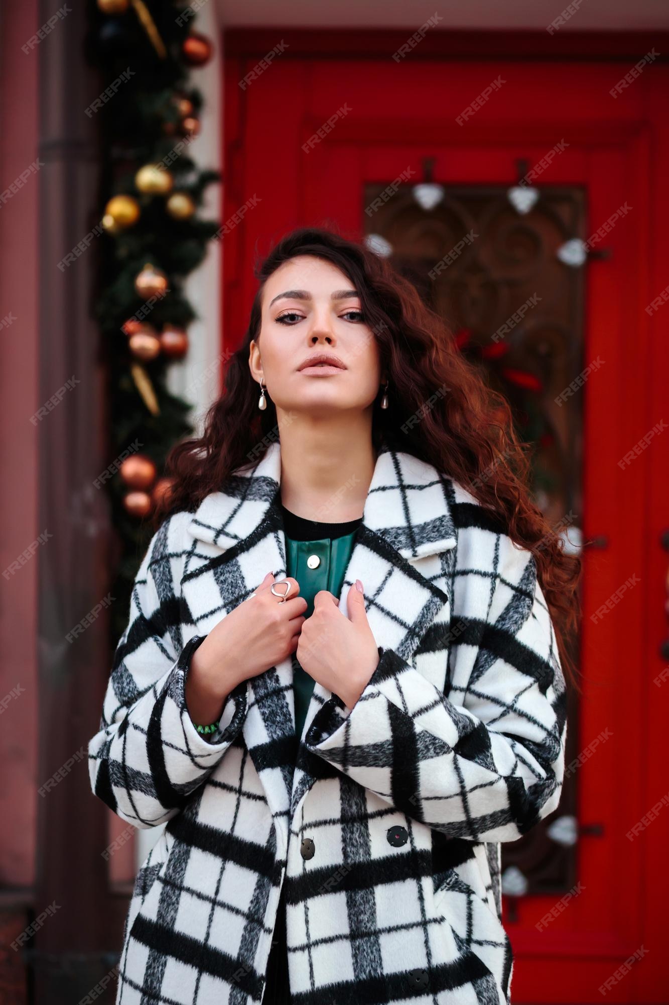 Mujer glamorosa en abrigo a cuadros blanco y negro frente a la puerta roja con adornos navideños, concepto de moda callejera de invierno. Foto Premium