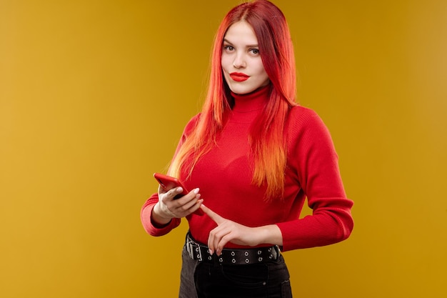 Mujer glamurosa con cabello rojo usando un teléfono inteligente con fondo amarillo