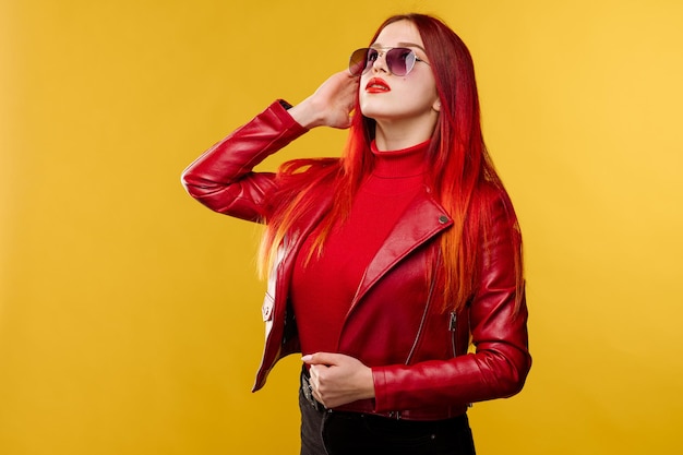 Mujer glamurosa con gafas de sol en una chaqueta de cuero roja y con el pelo rojo posando en el fondo del estudio