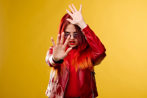 Mujer glamurosa con gafas de sol en una chaqueta de cuero roja y con el pelo rojo posando en el fondo del estudio