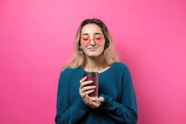 Mujer glamurosa con gafas en un suéter azul con un trago de café en un fondo rosa