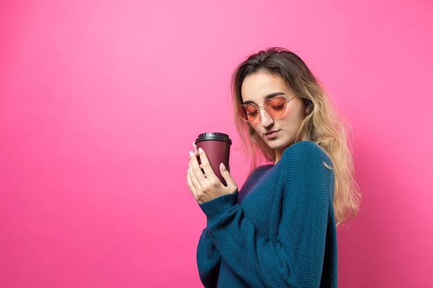 Mujer glamurosa con gafas en un suéter azul con un trago de café sobre un fondo rosax9