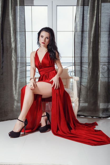 Mujer glamurosa sexy con cabello negro en un elegante vestido rojo sentada en un sillón en un dormitorio de lujo