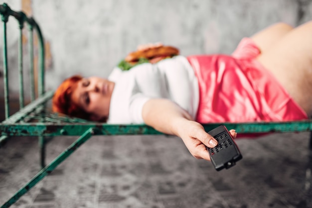 Mujer gorda con bocadillo en manos tumbado en la cama y ve la tele, pereza, bulímica y sobrepeso. Estilo de vida poco saludable, obesidad