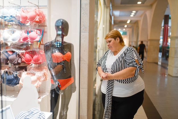 Mujer gorda cerca de la vitrina con ropa interior para cuerpo delgado. Persona de sexo femenino con sobrepeso soñando en la tienda con lencería, problema de obesidad