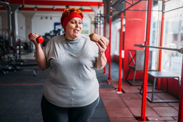 Mujer gorda con comida rápida y mancuernas en las manos, motivación, entrenamiento duro en el gimnasio. Concepto de quema de calorías, persona femenina obesa en gimnasio, quema de grasa, deporte contra alimentos poco saludables