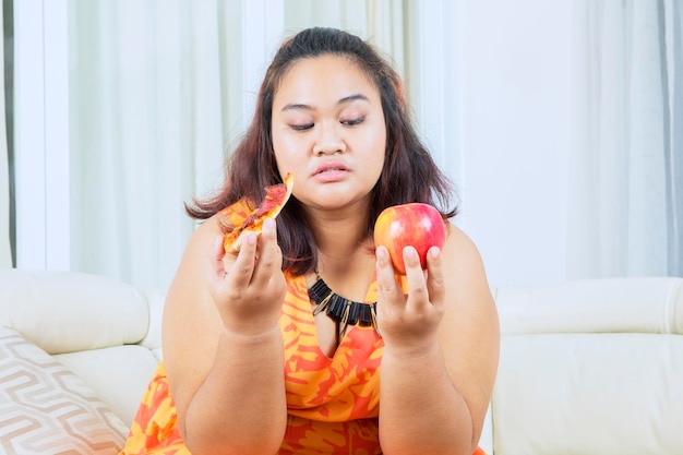 Mujer gorda confundida elige manzana o pizza en el sofá