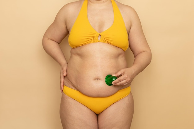 Mujer gorda en traje de baño amarillo masaje estómago grueso con frasco de vacío de cuerpo verde fondo beige Adelgazamiento lucha contra el sobrepeso obesidad y celulitis Personas de talla grande y concepto positivo del cuerpo