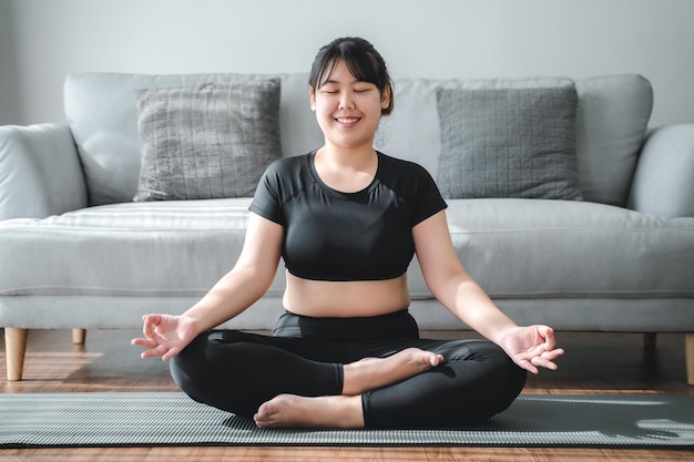 Mujer gordita asiática sentada en el suelo en la sala de estar practica la lección de yoga