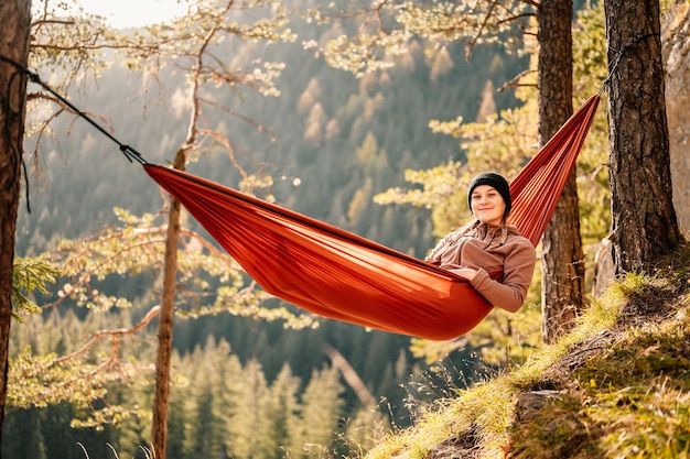 Mujer con gorra descansando en una cómoda hamaca durante la puesta de sol Relajándose en una hamaca naranja entre dos árboles de pino disfrutando de la vista