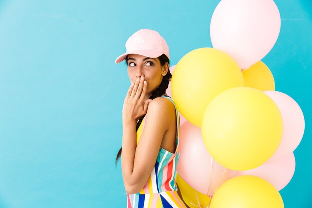 Mujer con gorra mirando a un lado en copyspace y coning su boca mientras sostiene globos de aire aislados en la pared azul