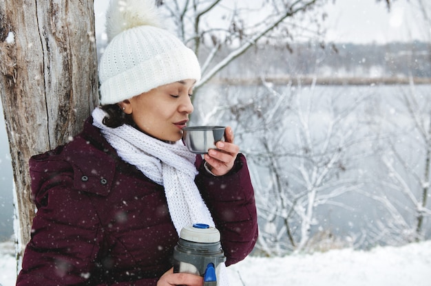 Mujer con gorro de lana de punto blanco disfrutando de un hermoso día de nieve mientras bebe té caliente. Nieve que cae